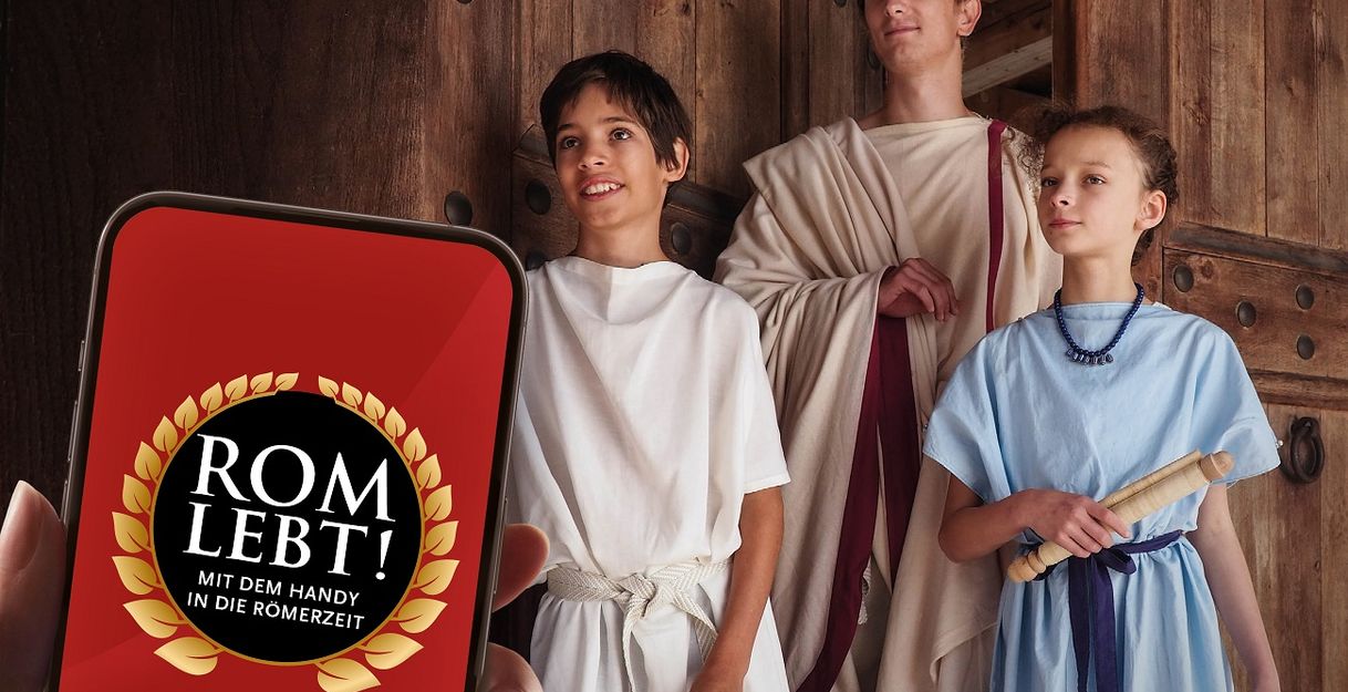 Drei als Römer der Antike gewandete Personen - ein Erwachsener und ein Junge und ein Mädchen- posieren in einer geöffneten Holztüre für ein Handy, das im Vordergrund sichtbar ist und den Namen der Ausstellung auf dem Display trägt
